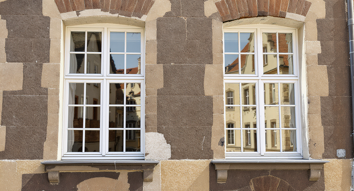 Zwei weiße Fenster mit Gittern sind in einem alten, verwitterten Steinmauerwerk eingelassen, wobei die Fenster reflektierende Oberflächen zeigen.